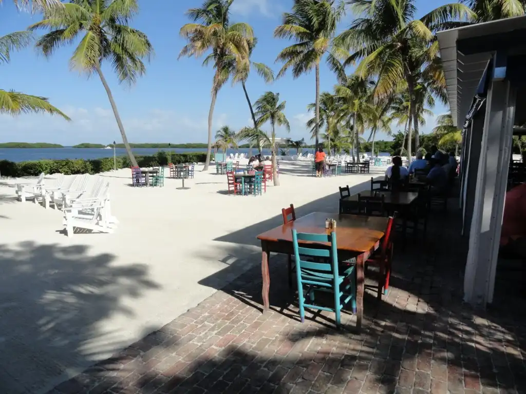 Outdoor tables on the sand under palm trees beside the water at Morada Bay Cafe, Islamorada, Florida